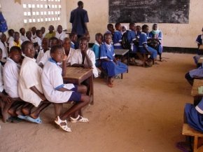 children sitting around tables on benches with a sandy floor