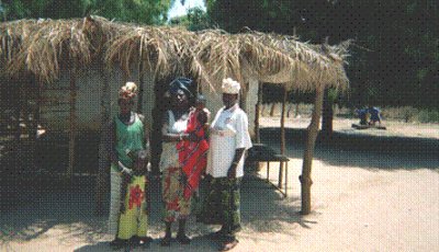 Bakalarr school cooks and their children outside the old kitchen which has a rustic veranda with a roof consisting of palm leaves