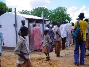 a toilet block is surrounded by a group of adults and children some of whom look inside