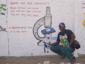 Kemo holds a microscope, in front of a large painting of a microscope